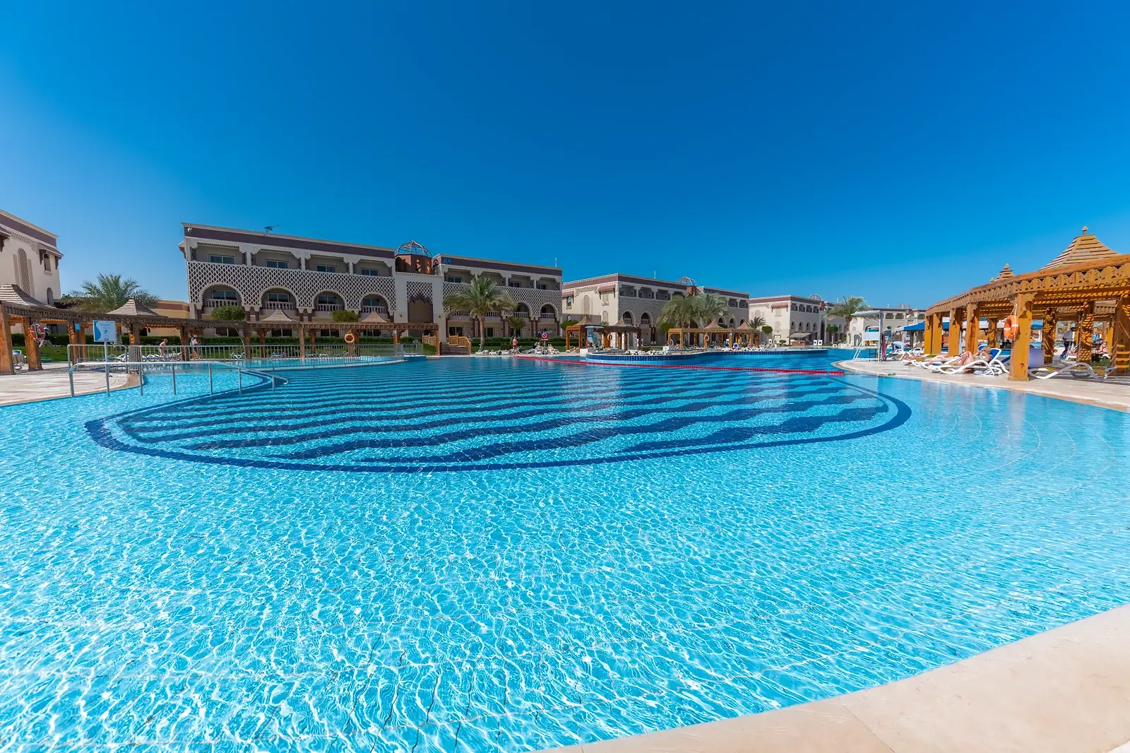 Groot zwembad met een golvend tegelpatroon in donkerblauw, ligbedden en een houten paviljoen voor de gebouwen met bogen van Sentido Mamlouk Palace Resort in Hurghada